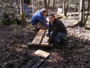 Two people are working in the woods, one cutting wood while the other smiles and assists with planks.