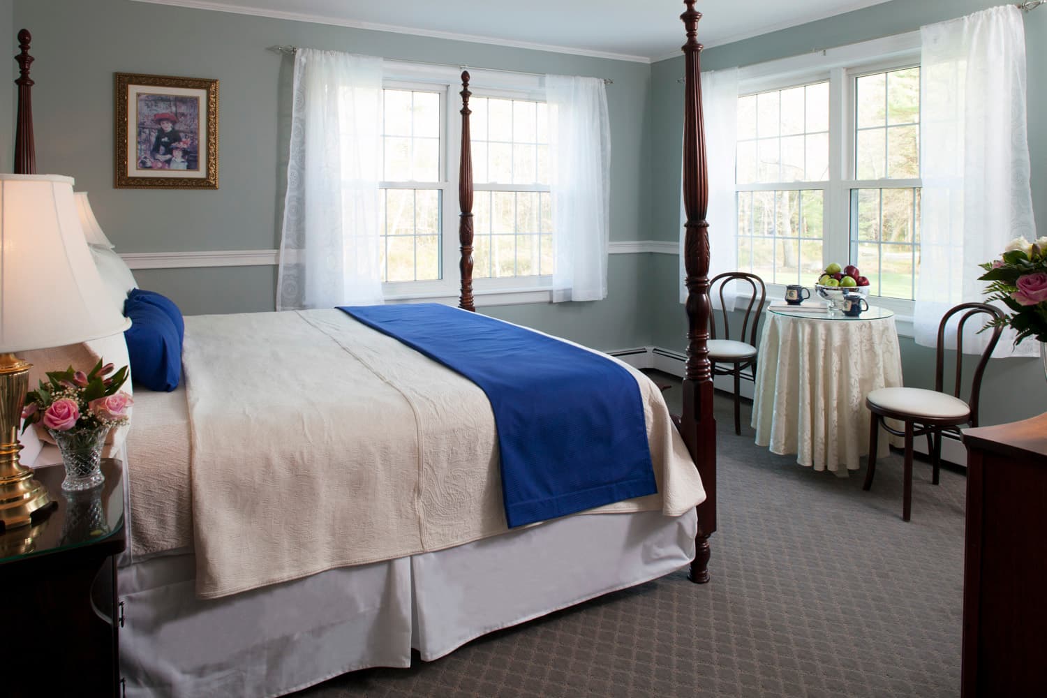 The king bed in the bedroom with corner windows and a small table and chairs set one of the windows.