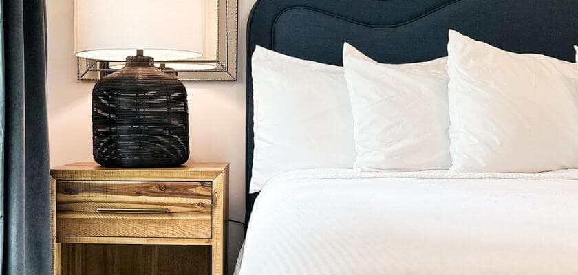 A cozy bedroom features a neatly made bed with white pillows and a wooden nightstand topped by a black lamp.