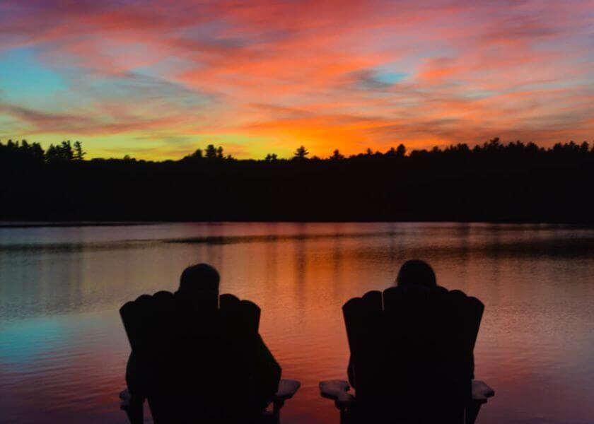 Silhouettes of two people sitting in chairs by a lake at sunset, with vibrant colors reflecting on the water.
