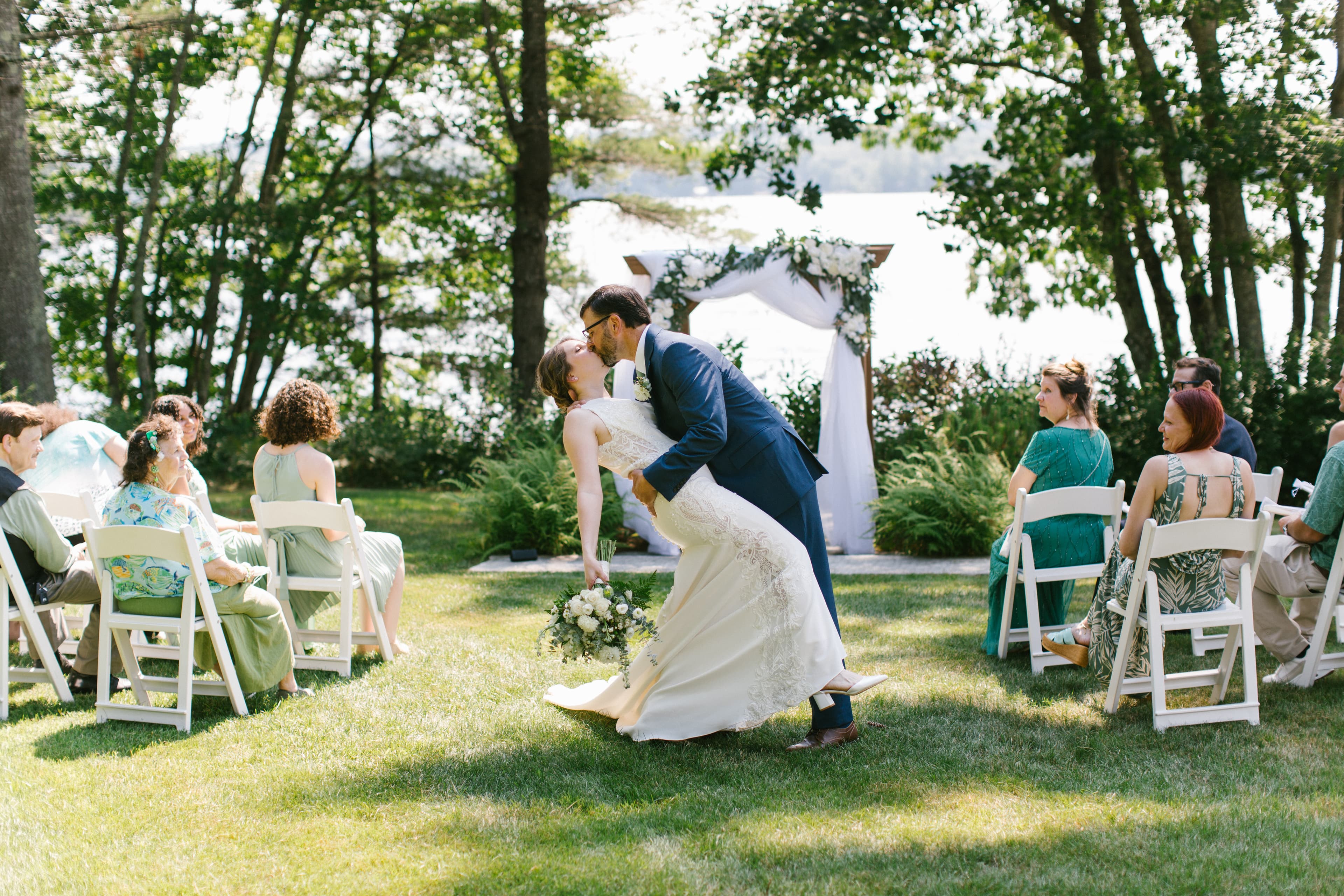 A couple shares a kiss during their outdoor wedding ceremony by a lake, with guests seated nearby.