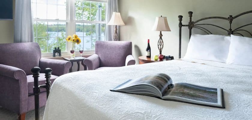 A cozy bedroom featuring a neatly made bed, two purple chairs, and a book open on the bed with a scenic view outside the window.