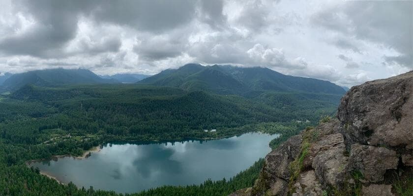 A panoramic view of a lake surrounded by dense forests and mountains under a cloudy sky.