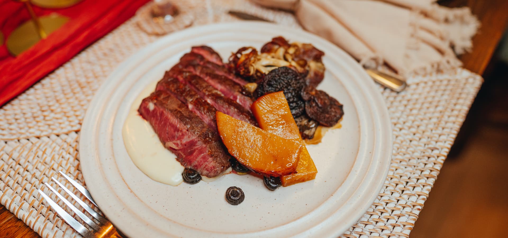 A plate of sliced beef next to roasted squash and a creamy sauce on a woven placemat.