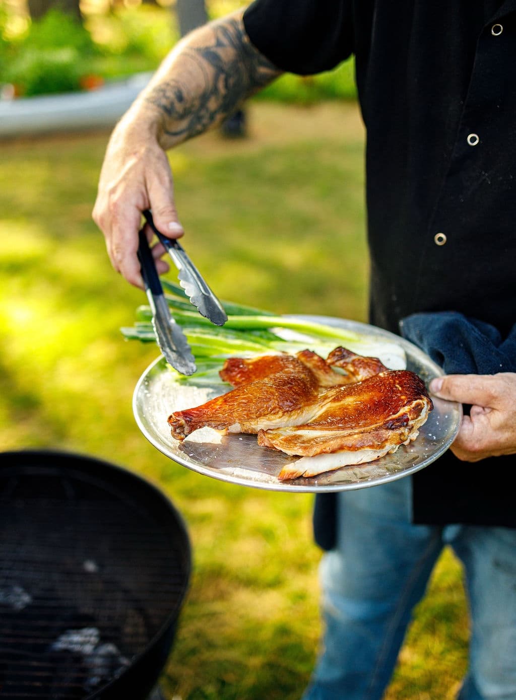 A person holds a plate of grilled chicken and green onions with tongs in a sunny outdoor setting.