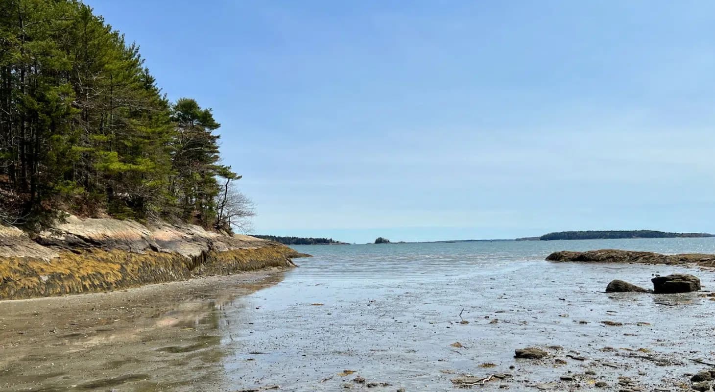 A serene coastal view with a sandy shore, gentle waves, and distant islands under a clear blue sky.