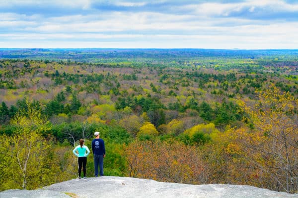 Two hikers stand on a rocky outcrop overlooking a vibrant green valley under a cloudy sky.