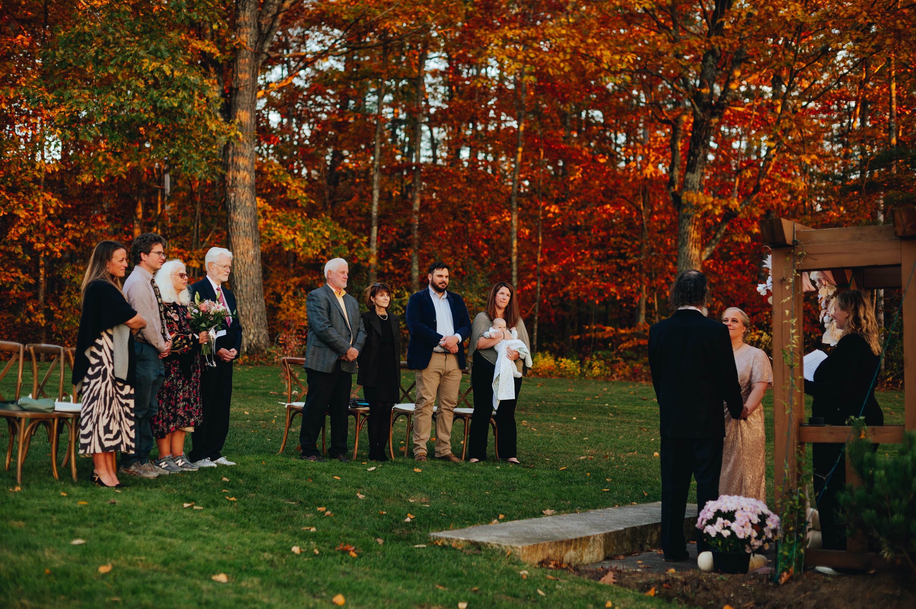 A wedding ceremony takes place outdoors with guests surrounded by vibrant autumn foliage.