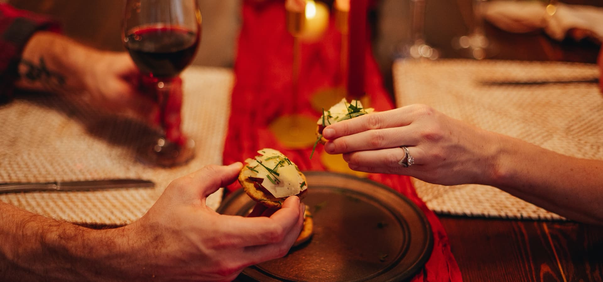 Two hands are reaching for appetizers on a plate, accompanied by a glass of red wine in a cozy dining setting.