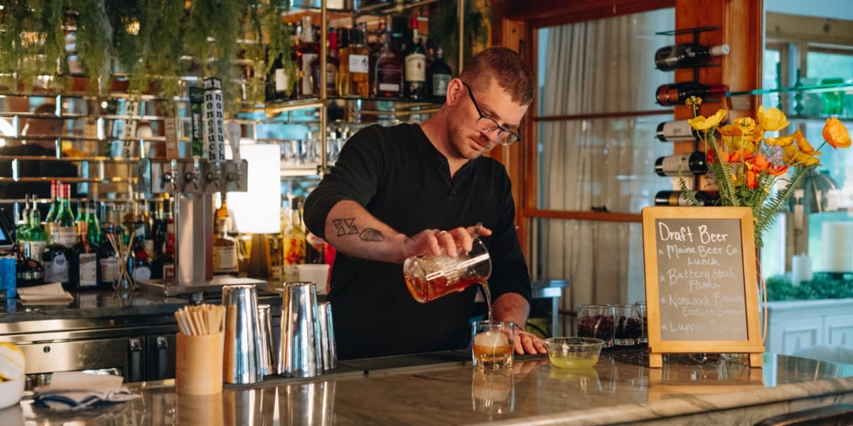 A bartender carefully pours a drink at a well-stocked bar adorned with flowers and a draft beer menu.