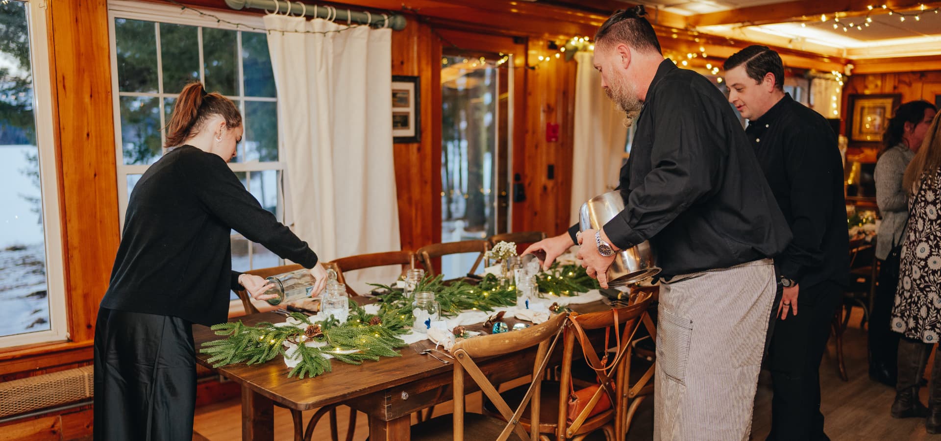 A group of people prepares a festive table with greenery and decorations in a cozy indoor setting.