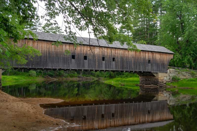 A wooden covered bridge spans a calm river, reflecting its image in the water.