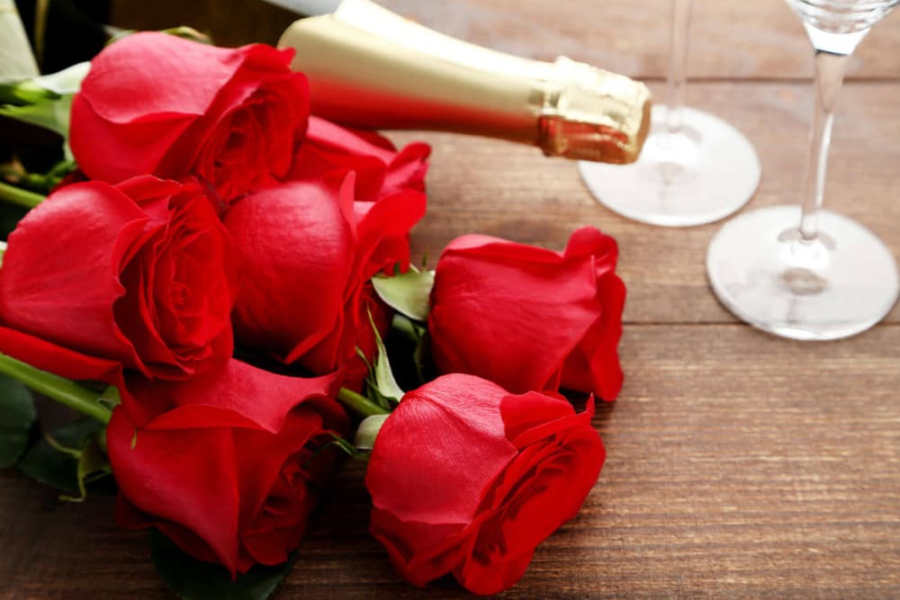 A bouquet of red roses beside a bottle of champagne and two glasses on a wooden surface.