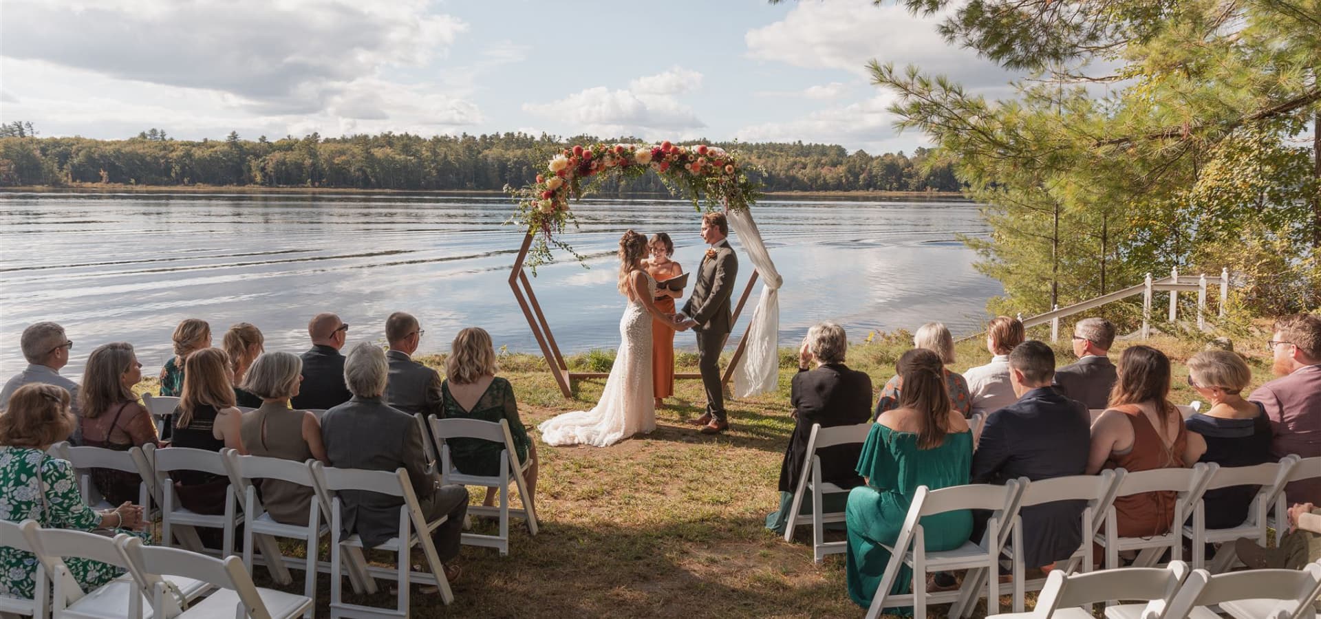 A bride and groom exchange vows under a floral arch by a serene lakeside, surrounded by seated guests.