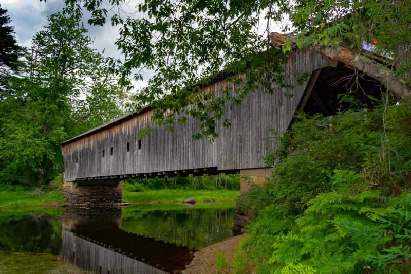 A rustic wooden covered bridge spans a tranquil stream surrounded by lush greenery.