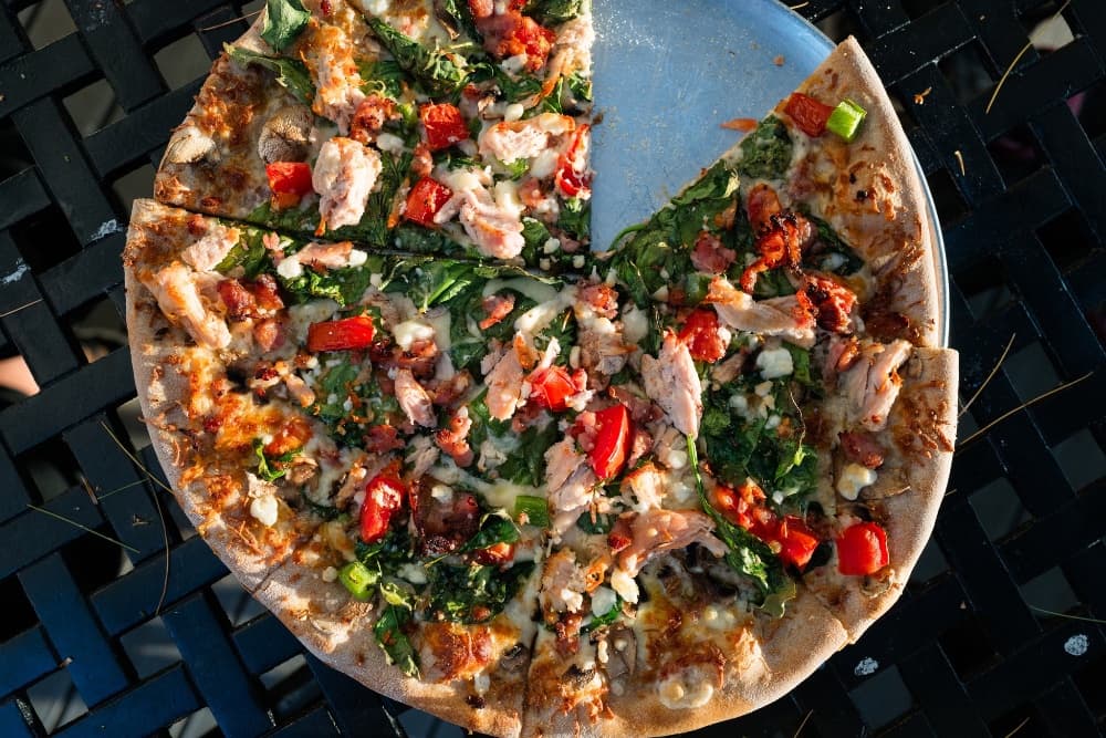 A partially eaten pizza topped with greens, tomatoes, and chunks of chicken on a metal plate.