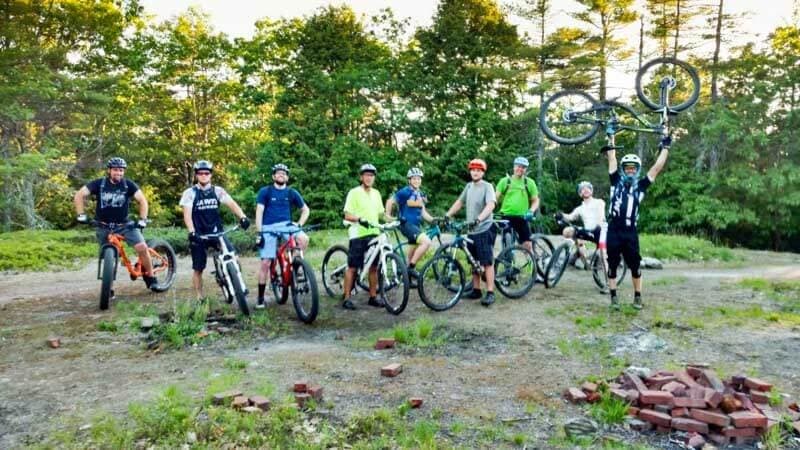 A group of eight mountain bikers poses outdoors, with one rider triumphantly lifting their bike above their head.