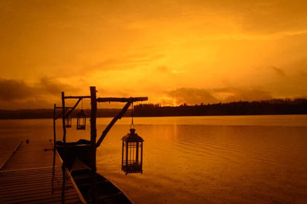 A lantern hangs from a wooden dock silhouetted against an orange sunset over a calm lake.