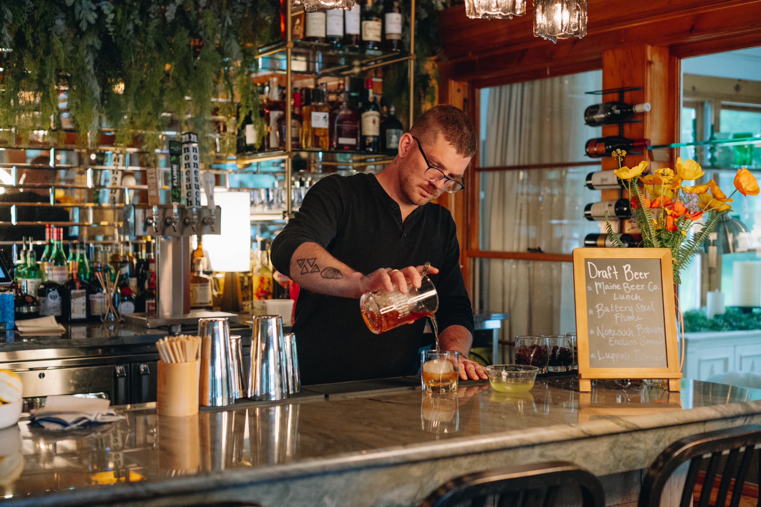 A bartender pours a drink at a stylish bar with an array of spirits visible in the background.