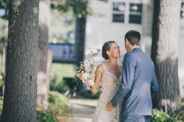 A bride in a wedding gown smiles at her groom while holding a bouquet in a serene outdoor setting.