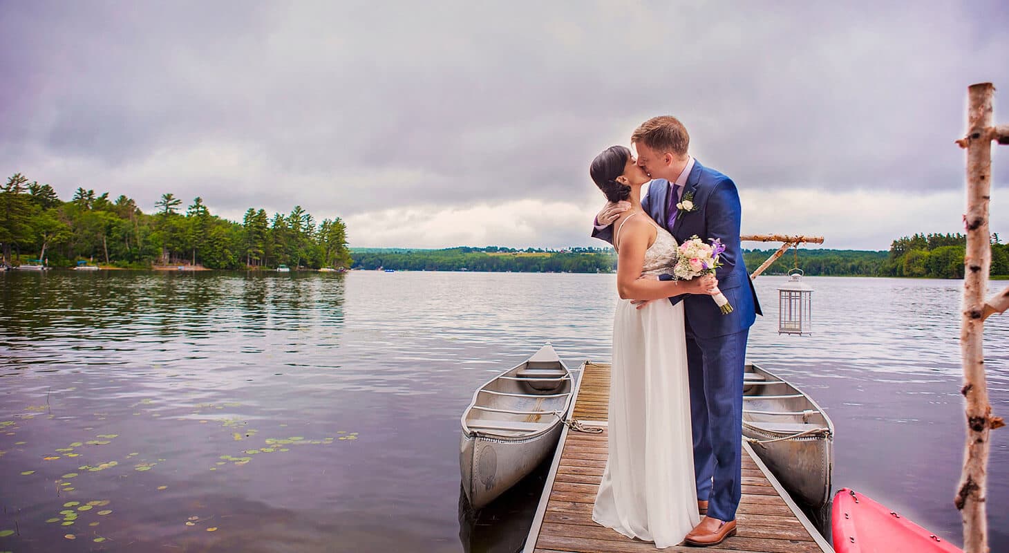 A bride and groom share a kiss on a dock by the water, surrounded by canoes and lush greenery.