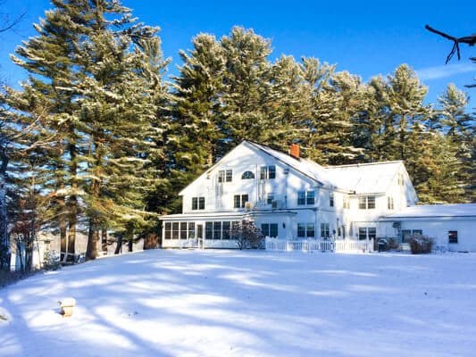 A white house surrounded by tall pine trees in a snowy landscape under a clear blue sky.