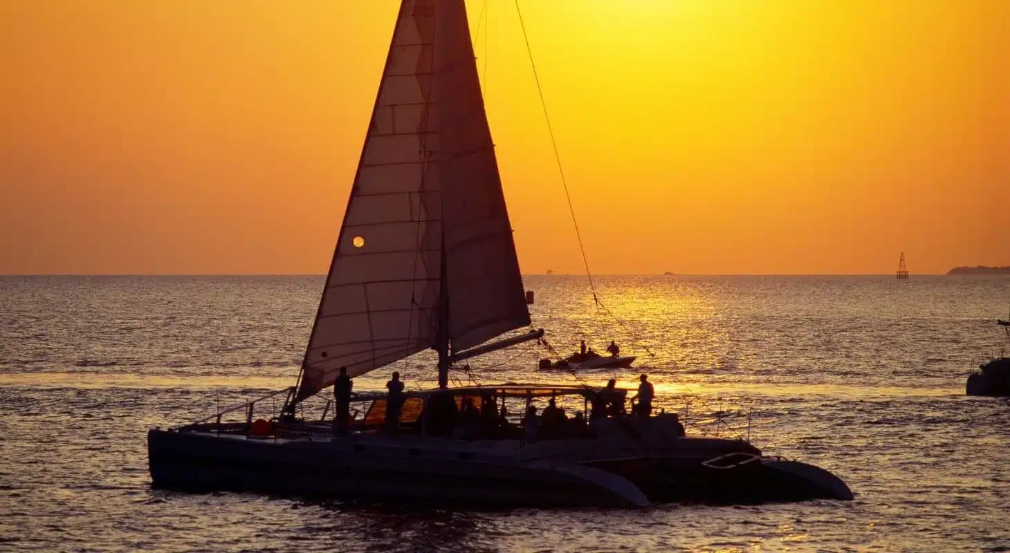 A silhouette of a sailing catamaran against a vibrant orange sunset over the ocean.