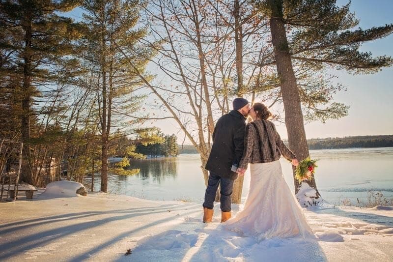 A couple kisses in a snowy landscape near a lake while surrounded by trees.