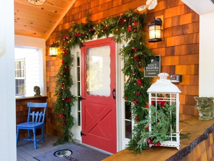 A red door adorned with greenery and festive decorations opens into a cozy entryway with a blue chair and a lantern.