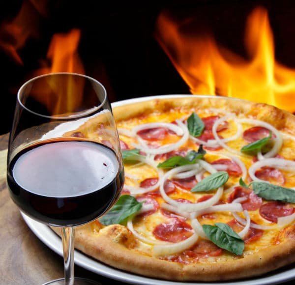 A glass of red wine next to a pepperoni pizza topped with onions and basil, with flames in the background.
