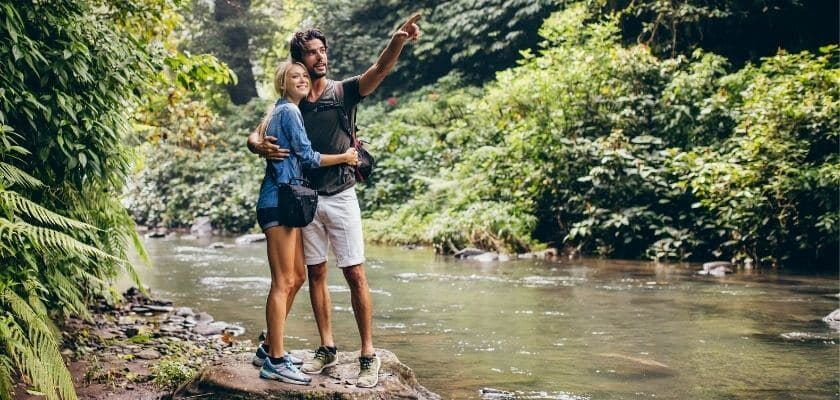 A couple hugs by a river, with the man pointing toward the trees.
