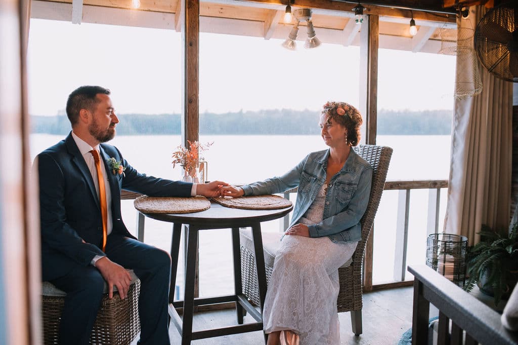 A couple sits at a table by a lake, holding hands and gazing at each other.