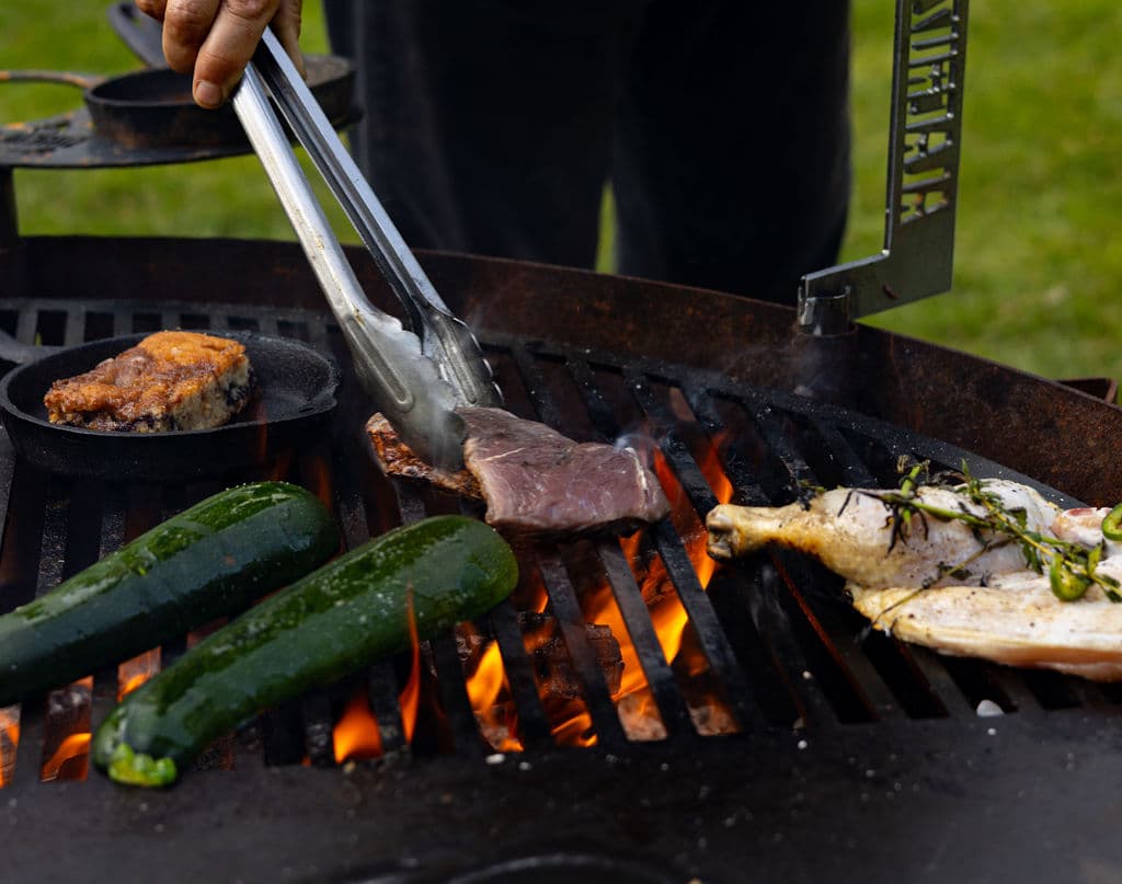 A person uses tongs to flip meat and vegetables on a grill with flames.