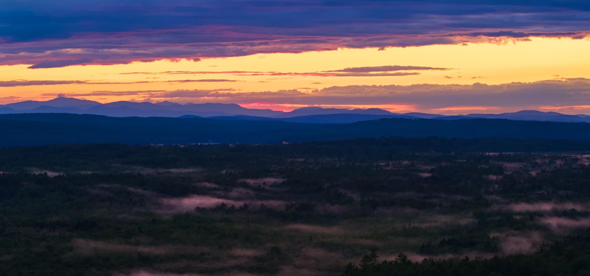 A vibrant sunset casts warm hues over misty valleys and distant mountains.
