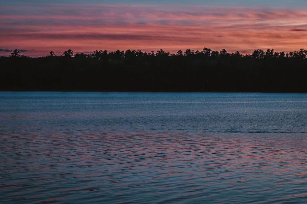 Calm lake at dusk with a colorful sky and silhouette of trees.