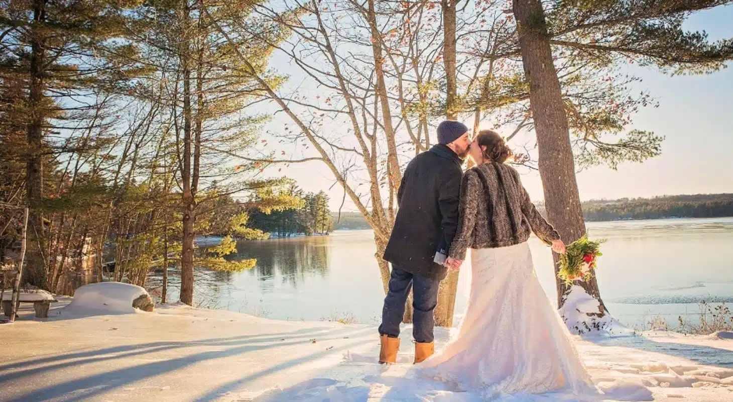 A couple shares a kiss in a snowy landscape by a lake, surrounded by trees.