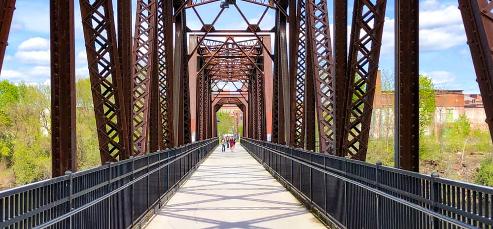 A pedestrian bridge with a metal frame stretches into the distance, flanked by greenery under a partly cloudy sky. A pedestrian bridge with a metal frame stretches into the distance, flanked by greenery under a partly cloudy sky.