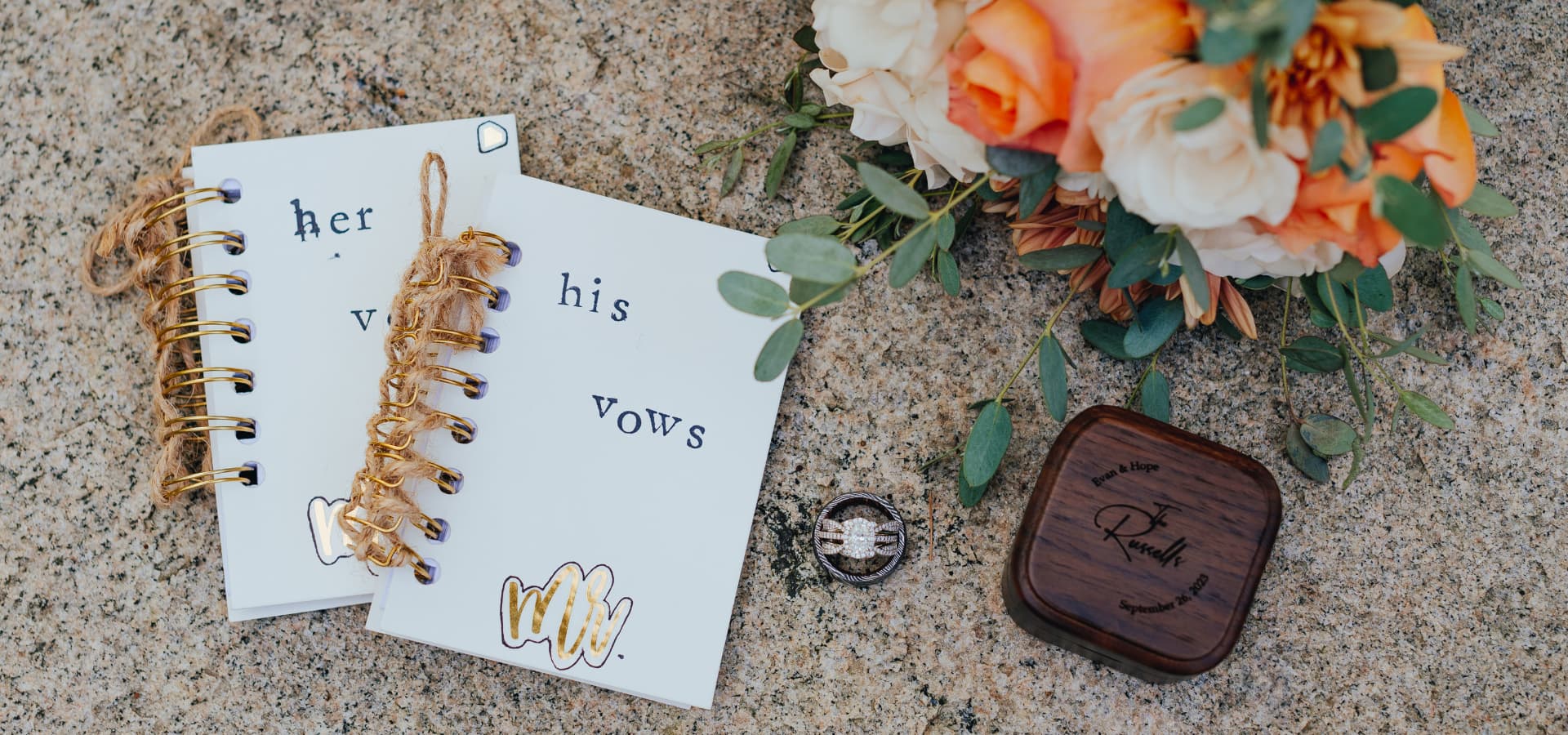 A flat lay featuring wedding vow books labeled "his" and "her," a wooden ring box, an engagement ring, and a floral arrangement.