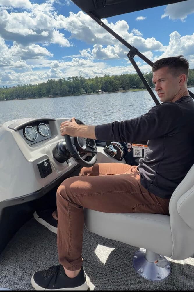 A man steers a boat while sitting at the helm, surrounded by water and a blue sky.