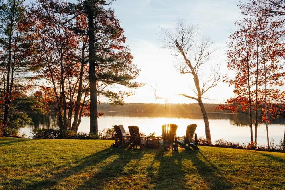 A serene lakeside scene at sunset with four empty chairs and colorful autumn foliage.