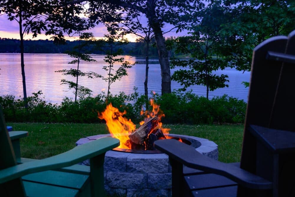 A bonfire burns in a stone pit beside a calm lake at sunset, framed by green chairs.