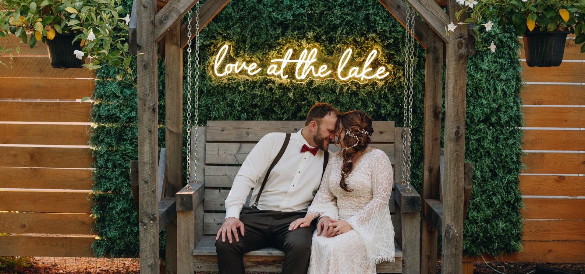 A couple in formal attire sits together on a wooden swing under a green wall, with a neon sign reading "Love at the lake" above them.