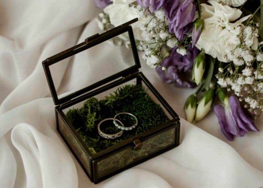 Two wedding rings nestled in a glass box with moss, beside a bouquet of purple and white flowers. Two wedding rings nestled in a glass box with moss, beside a bouquet of purple and white flowers.