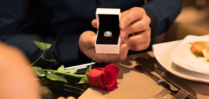 A person holds an open ring box alongside a red rose and a plate of food.