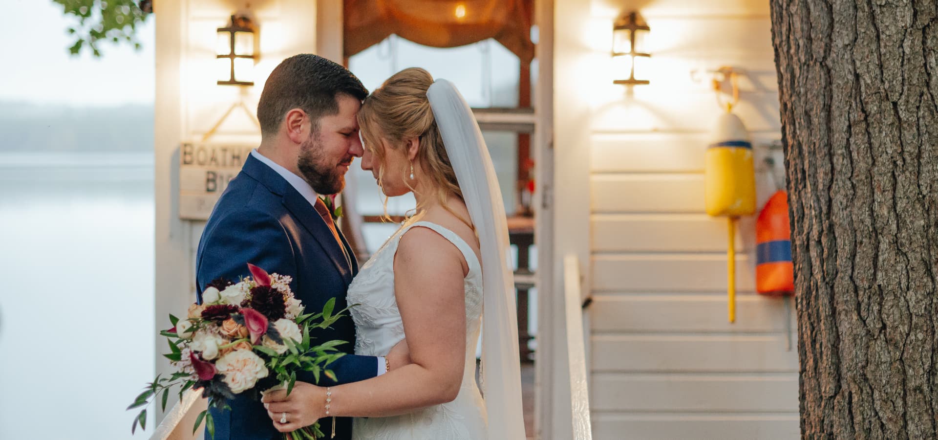 A bride and groom embrace lovingly in front of a lakeside boathouse.