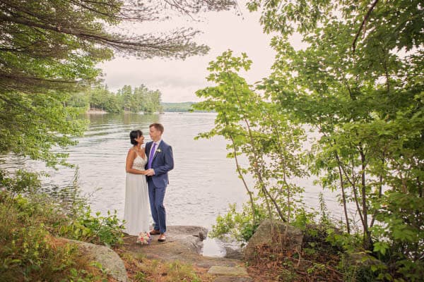A couple in wedding attire stands together on a rocky shore, surrounded by trees and a calm lake.
