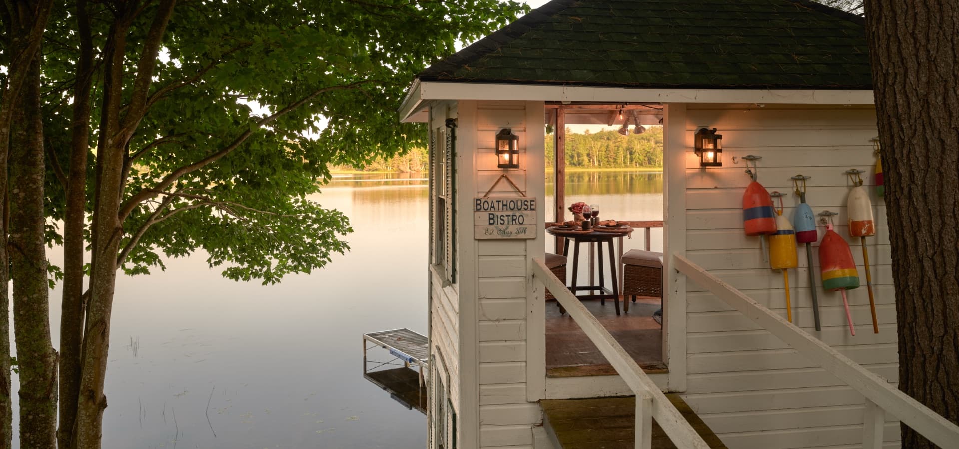 A charming boathouse bistro with a view of a calm lake, surrounded by trees and decorative buoys.