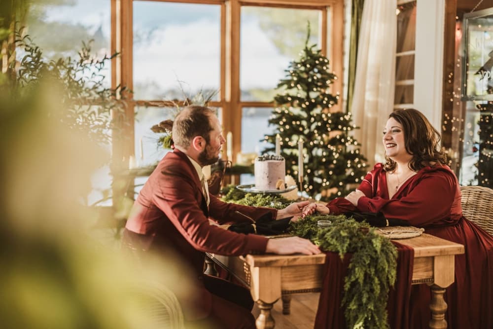 A couple sits at a cozy table decorated with greenery, smiling at each other in a festive setting with a Christmas tree.