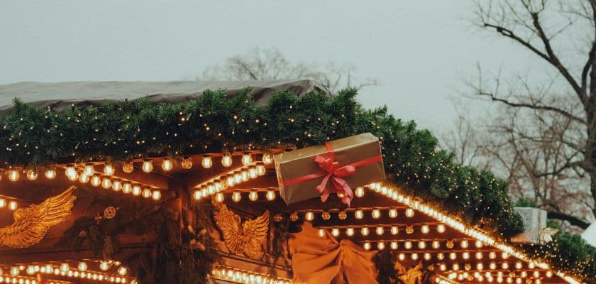 A festive gift box with a red ribbon hangs from a decorated carousel, surrounded by twinkling lights and greenery.