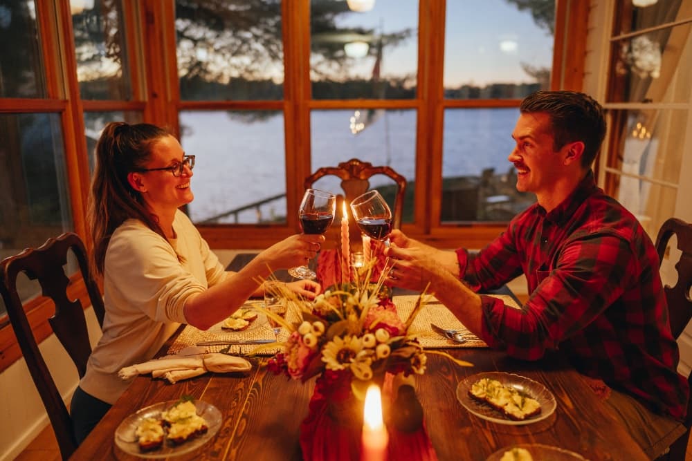 A couple toasts with red wine at a cozy dinner table by the water at sunset.